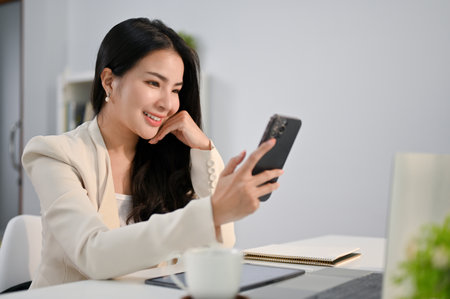 Beautiful millennial Asian businesswoman hand on chin, looking at her phone screen, using her phone, scrolling on social media or using mobile app at her desk.の写真素材