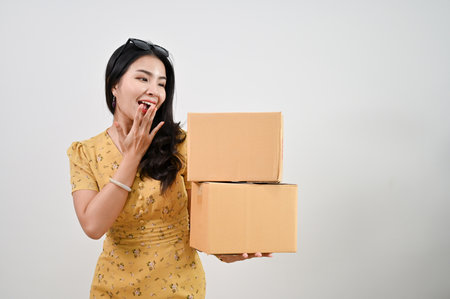 Shocked and excited young Asian woman in casual clothes covering her mouth and holding cardboard boxes while standing against an isolated white background. shipping, online shopping conceptの写真素材