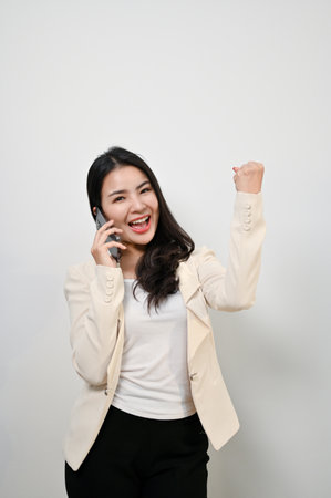 Portrait, Cheerful millennial Asian businesswoman talking on the phone with joy and happiness, celebrating success, rejoicing, yes gesture, standing against an isolated white background.の写真素材