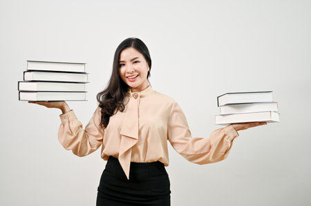 Smiling millennial Asian female teacher holding a stack of books while standing against an isolated white background.の写真素材