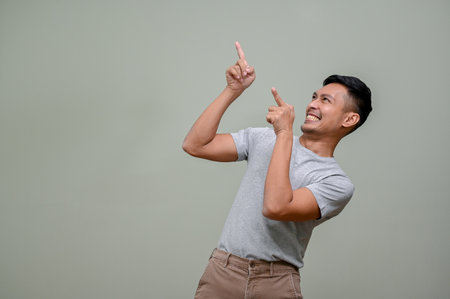 Cheerful and smiling millennial Asian man in casual clothes looking aside and pointing his fingers up at the empty space while standing against an isolated green background. hot promotion, discountの写真素材