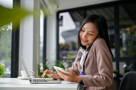 Attractive and professional millennial Asian businesswoman or female accountant is on the phone with her business client while using calculator and working at her desk in the office.の写真素材