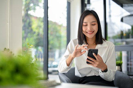 Smiling and happy millennial Asian businesswoman chatting with her friends on her phone, scrolling on social media or watching video on the internet, using her phone while relaxing in the office.の写真素材