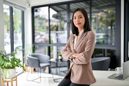 Confident and successful millennial Asian businesswoman or female CEO in formal business suit sits on table in her modern office with crossed arms.の写真素材