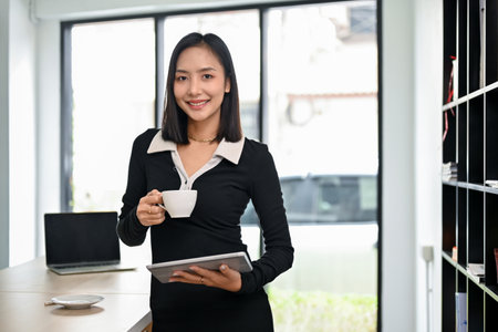 Confident and elegant millennial Asian businesswoman or female boss in trendy clothes, holding a coffee cup and a tablet, standing in her office.の写真素材