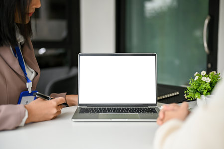 Two professional millennial Asian businesswomen discussing and working together in the meeting room, with a laptop white screen mockup on table.の写真素材