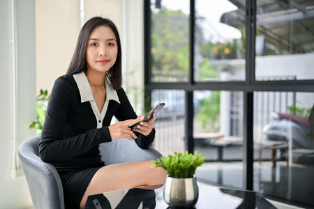 Attractive millennial Asian businesswoman in casual black dress holding her phone and looking at the...
