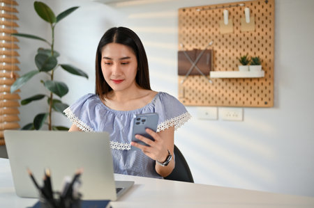 Charming young Asian female office worker in casual clothes using her phone and laptop in her office.の写真素材