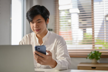 Handsome and smart millennial asian businessman or male business consultant using his phone and laptop at his desk.の写真素材