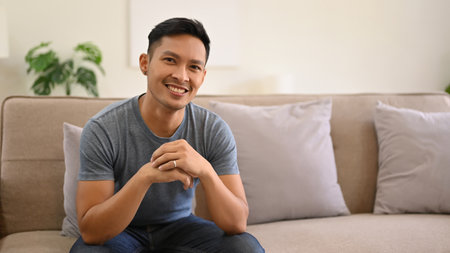 Smiling and handsome millennial Asian man in casual clothes sits on a comfy sofa in his minimal living room. lifestyle conceptの写真素材