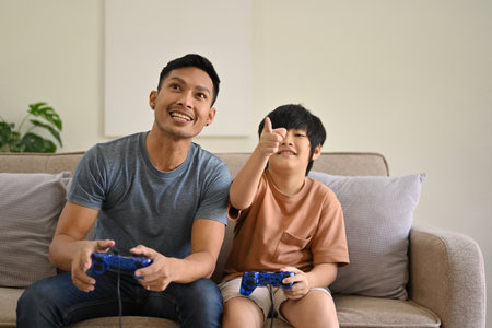 Joyful and excited Asian dad and son with joysticks are playing video games, competing in online kid's games, and spending fun time at home together.の写真素材