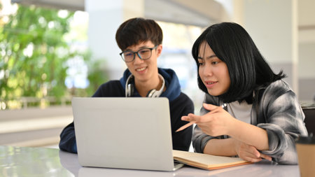 Two smart and happy young Asian college students are preparing for school exam or doing a schoolwork on laptop together at the campus building.の写真素材