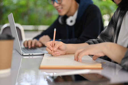Cropped image of two Asian young college students doing their school project together, using laptop and writing something on paper.の写真素材