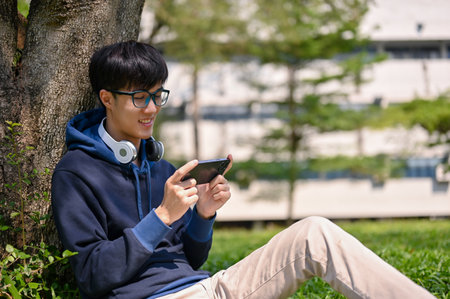 Portrait of a happy young Asian male college student using his smartphone or playing mobile games while sitting under the tree in the campus park.の写真素材