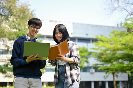 Happy young Asian female and male college student stands with a laptop and school book in front of their campus building.の写真素材