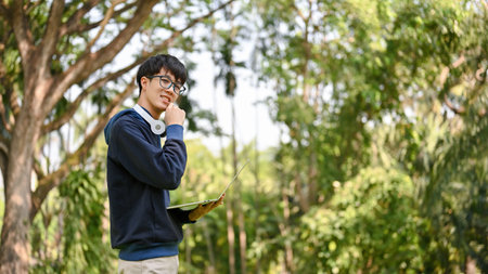 Portrait of a handsome smart young Asian male college student with his laptop standing in the greenery park.の写真素材