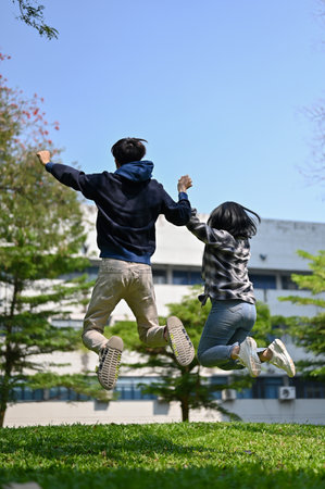 Carefree and overjoyed Asian college students are holding hands and jumping to celebrate their school life success together at the campus park. back viewの写真素材