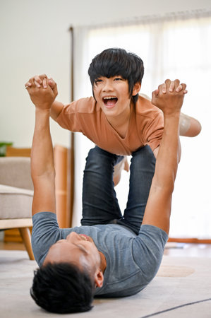 Loving Asian dad lying on the living room floor playing airplane with little son, lifting up his son in the air, spending fun family time together.の写真素材