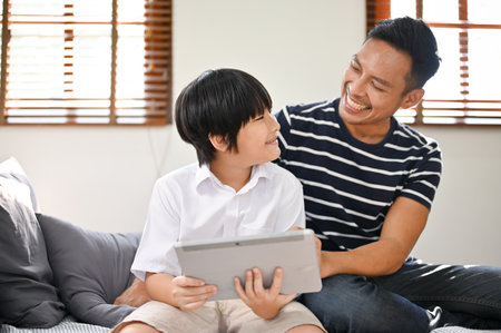 Joyful and happy Asian dad enjoys playing online kid's games or watching kid's video content on a tablet with his son in the living room.Â leisure and family conceptの写真素材