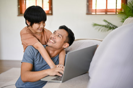 Cute little Asian boy hugging his father from behind while he works on his laptop in the living room.Â happy family concepttの写真素材