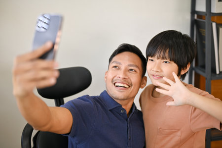 Cheerful and smiling Asian son and dad are taking a picture together with smartphone at home. taking selfie, happy family leisure timeの写真素材