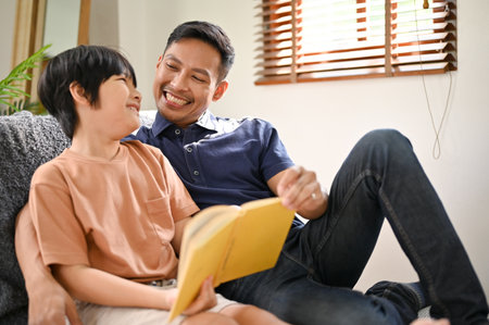 Happy and cheerful Asian dad reading fairytale and having a good time with his little son on the weekend at home.の写真素材