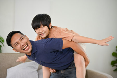 Smiling and laughing Asian dad playing with his son, piggyback, having a fun family time together at home.の写真素材