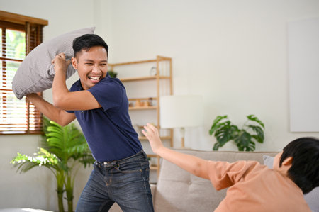 Playful and happy Asian dad enjoying playing pillow fight with his little son in the living room, having fun family time on the weekend together. happy family leisure conceptの写真素材