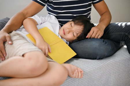 Relaxed and happy young Asian son listened to a fairytale and fell asleep on his father's lap on the sofa in the living room.の写真素材