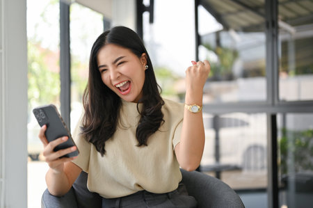 Overjoyed and excited millennial Asian woman screaming with joy, looking at her phone screen, rejoicing, receiving good news, winning lottery.の写真素材