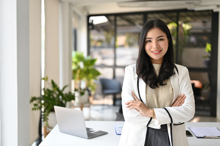Confident and successful millennial Asian businesswoman or female CEO in white suit stands with arms crossed in the office.の写真素材