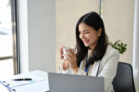 Charming and happy millennial Asian businesswoman or female marketing manager enjoys having morning coffee at her desk.の写真素材