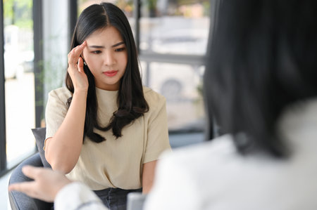 Stressed and thoughtful millennial Asian woman feeling dizzy and headache while having a serious talk with her personal financial consultant.の写真素材