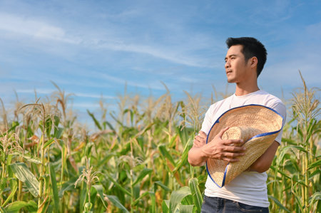 Successful millennial Asian male farmer or corn farm owner with his straw hat in his hand stands with arms crossed in his cornfield.の写真素材