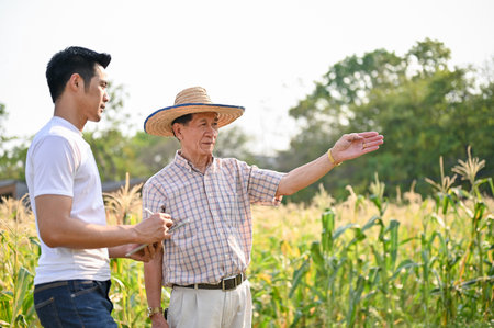 An Asian farmer in a straw hat walking and talking with a male supplier in a cornfield during a corn quality inspection.の写真素材