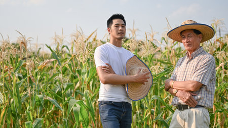A successful millennial Asian male farm owner or supplier and an elderly farmer are standing with arms crossed in a cornfield.の写真素材