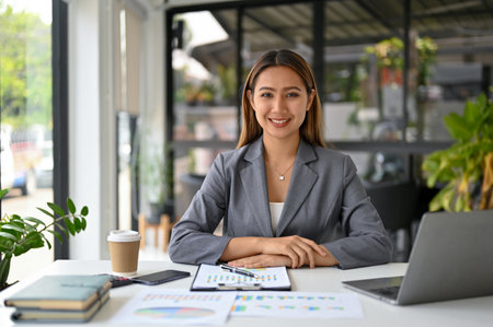 Attractive and confident millennial Asian businesswoman or female boss in formal business suit sits at her desk in modern office.の写真素材