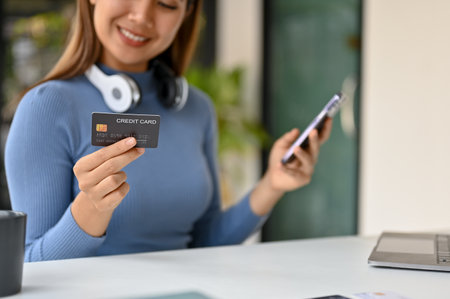 Close-up image of a smiling millennial Asian woman sits at a table, holds her credit card and smartphone, and uses mobile banking app to pay online bills. cashless payment, e-commerce, e-wallet.の写真素材