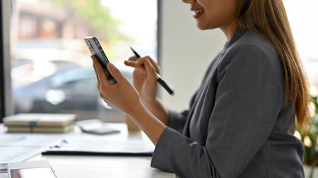 Close-up side view image of an elegant millennial Asian businesswoman or female secretary using her smartphone at her desk. text, chat, message, sms, mobile application, online shopping.の写真素材