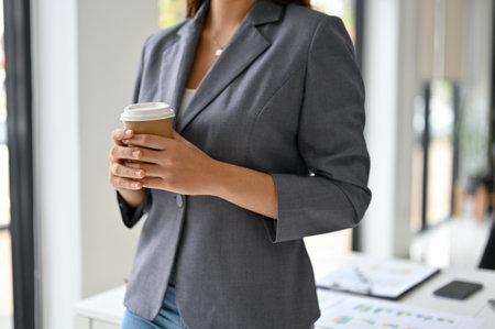 Cropped image of a gorgeous and confident Asian female CEO or boss in formal business suit stands in her office and holds a takeaway coffee cup.の写真素材