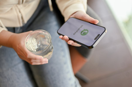 Chiang Mai, Thailand - March 31 2023: A female sipping water, relaxing in a cafe and using WhatsApp on her phone. iPhone with WhatsApp logo on screen.のeditorial素材