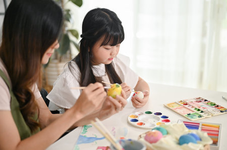 A cute young Asian girl focuses on painting Easter eggs with watercolors, spending a wonderful time with her mother at home.の写真素材