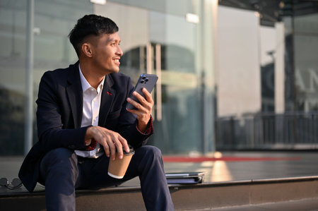 Attractive and happy millennial Asian businessman or male worker in formal business suit using his smartphone and sipping coffee while sitting on stairs outside of the building.の写真素材