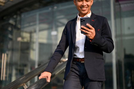 Handsome and successful millennial Asian businessman in a formal business suit holding his smartphone while walking down the stairs in a city. cropped imageの写真素材