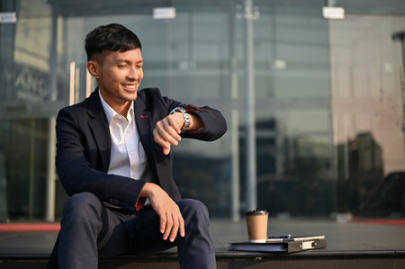 Happy and smiling millennial Asian businessman checks the time, looking at his watch, while sitting on the stairs in front of the company building.の写真素材