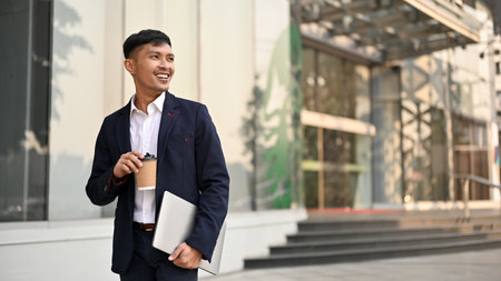 Confident and successful millennial Asian businessman in a formal business suit walking in a city with his takeaway coffee and laptop. Urban lifestyle conceptの写真素材
