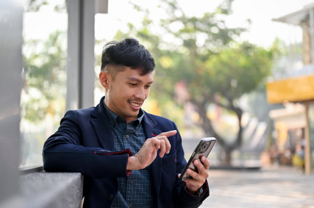 Happy and handsome millennial Asian businessman using his smartphone while waiting for a bus or taxi in the city.の写真素材
