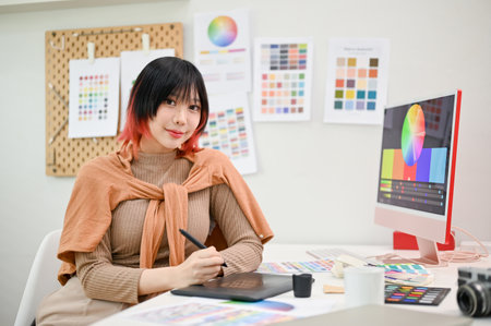 Attractive and smiling young Asian female graphic designer or interior designer sits at her desk in her studio.の写真素材
