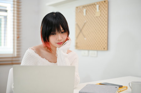 Thoughtful young Asian woman focusing on her tasks on laptop, browsing internet, reading online...