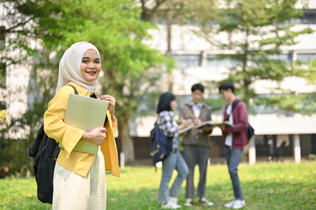 Attractive and smiling young Asian Muslim female college student in hijab carrying her laptop and...